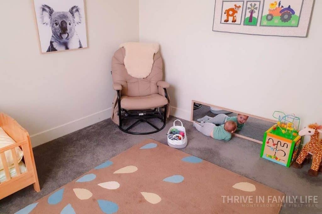 Montessori nursery with Montessori 4 month old shelf space on the floor with a baby on floor in front of floor mirror with basket of toys behind him and a rocking chair in the corner. 