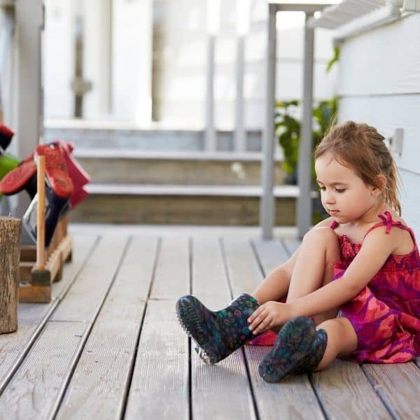 young toddler girl in pink dress putting on her own rain boots on front porch