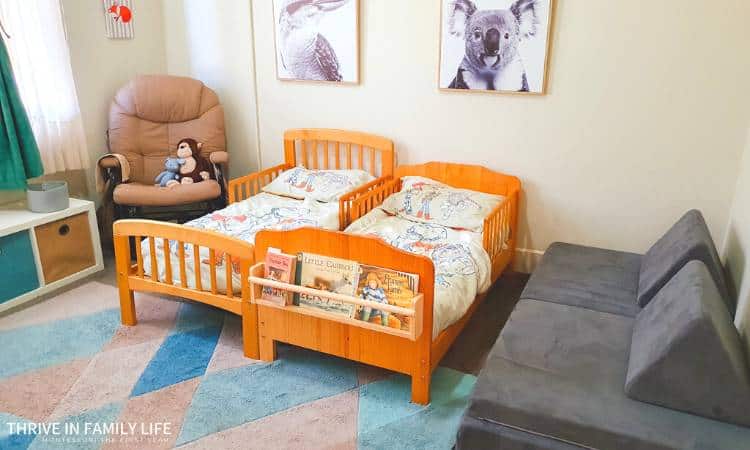 montessori shared bedroom for two boys with two wodden toddler beds, rocking chair, small shelf with blue & beige drawers, and grey kids couch nugget. blue rug on floor.
