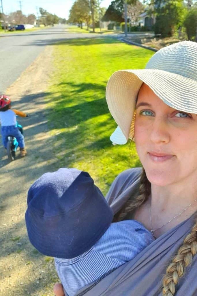 Woman baby wearing 2 month old baby outside while 2 year old toddler rides blue balance bike.