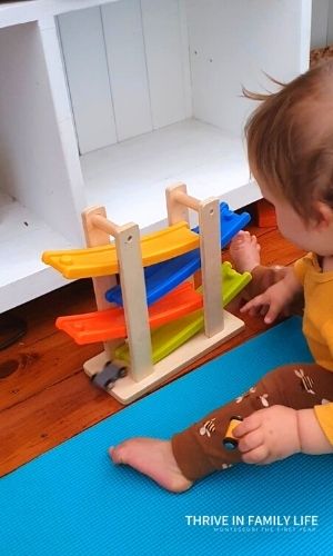 12 month old in yellow shirt and brown pants putting wooden grey & yellow cars on tracking toy. 