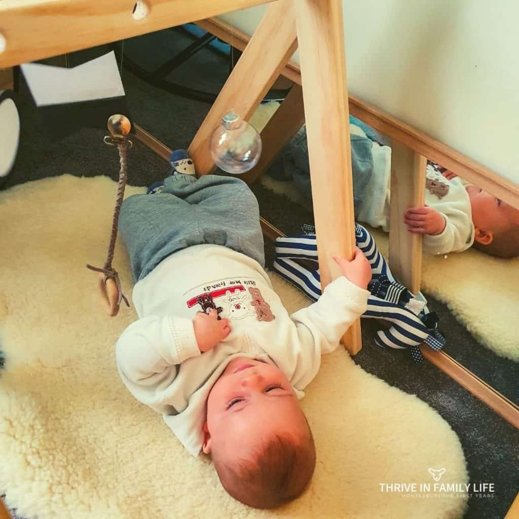 DIY Montessori Mobile for 4 month old laying on a white sheepskin rug next to a floor mirror. 