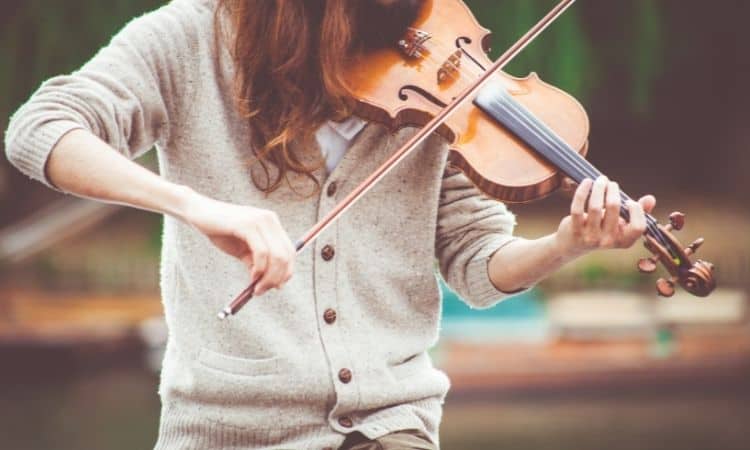 Woman in beige sweater and brown hair outside playing a violin