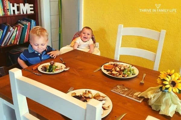 Montessori meal time at wooden table with white chairs with toddler in tripp trapp chair and smiling 4 month old baby in high chair watching. 