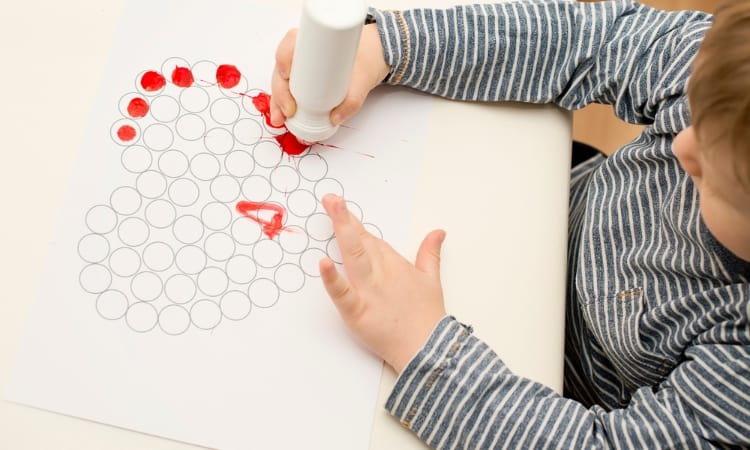 toddler boy in blue & white striped shirt sitting at table using red dabber on empty circles in the shape of a heart. 