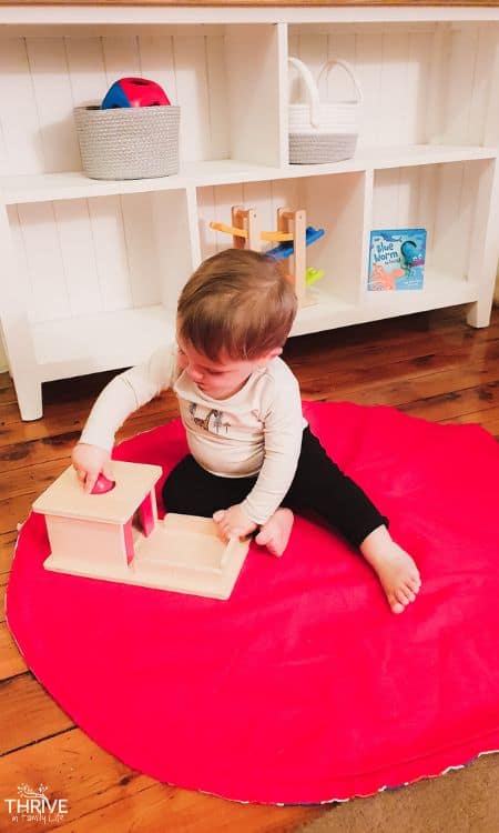Montessori toys 15 months - little toddler on red circle mat using a Montessori object permanence box.