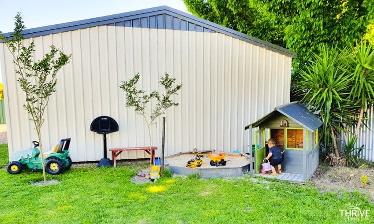 Outdoor play area for toddler - 15 months old. Small cubby house painted grey with green trim, small octagon sandbox, green tractor pedal toy, and toddler sized black basketball hoop. All in front of a beige shed wall. 